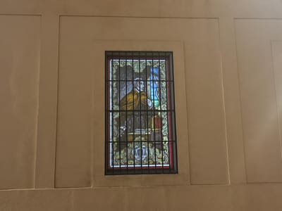 The image captures a stained-glass window set within a recessed panel in a wall. The wall is beige. The window features a figure, likely a historical or religious person. The person has white hair and is wearing a gold and gray cloak, with a white or light-colored shirt and black trousers. There is blue glass on the upper right side of the figure. The window is adorned with an ornate design along its borders, in gold and red. The window is protected by a black metal grid. The recessed panel in the wall is flanked by vertical trim on either side. The location of the window is in the city of Córdoba, Spain.