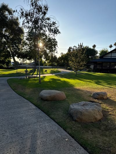 The image depicts a sun-drenched park in Glendale, USA, with a paved pathway curving through a lush green lawn. Large, weathered rocks are strategically placed on the grass, and a Japanese-style pagoda-like building with a dark roof and intricate wooden trim stands to the right, partially obscured by trees. Sunlight filters through the leaves of tall trees, casting long shadows across the manicured landscape. A clear blue sky stretches above, with a few palm trees visible in the distance, hinting at the Californian locale.