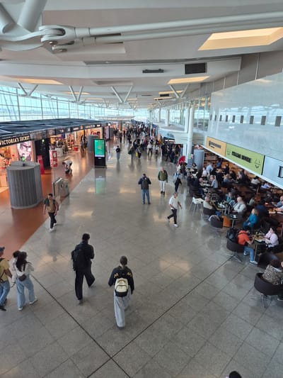 The image depicts an indoor scene at what appears to be an airport terminal in Moreira, Portugal. The perspective is angled, showing an overview of the space. Many people are present, engaged in various activities such as walking, sitting at a cafe, and browsing in shops. The 'Italian House' cafe is visible with customers seated at tables. Several retail stores are present, including a 'Victoria's Secret' location. The architecture features a high ceiling with prominent lighting fixtures and glass windows. The floor is tiled and predominantly gray. The image captures a bustling atmosphere with a concentration of people in the cafe area and along the walkway.
