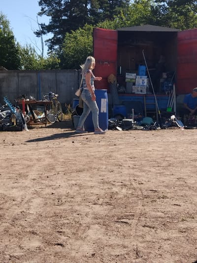 The image depicts an outdoor scene in Irshansk, Ukraine, likely a junkyard or a makeshift marketplace. In the foreground, a woman with blonde hair, wearing a patterned top and light-wash jeans, walks away from the camera towards a red trailer. She has a white crossbody bag. The ground is composed of dirt and gravel. Behind her, various items are strewn about, including a blue plastic barrel, a bicycle on a stand, and piles of scrap metal and other miscellaneous objects. To the left, a concrete wall is visible, with trees and foliage beyond it. The red trailer, open at the back, is filled with boxes, containers, and more assorted items. A person in a blue shirt sits in the right background, facing away from the camera. The sky above is clear and blue, suggesting a sunny day.