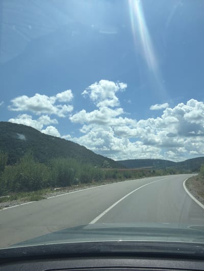 The image depicts a road curving through a hilly landscape under a bright, sunny sky with scattered cumulus clouds. The foreground shows the dashboard and windshield of a vehicle, with a reflection of the sky and clouds on the glass. The road is paved and appears to be a two-lane highway with a white line marking the center. On the left side of the road, there is a lush green hillside covered with trees and bushes. On the right side, the landscape continues with rolling hills and vegetation. The overall scene suggests a scenic drive through a rural area. This image is taken in the vicinity of the village of Garchinovo, Bulgaria.