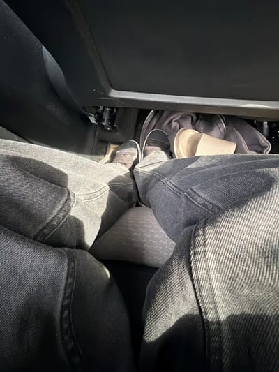 The image depicts a close-up, overhead view of a person's legs and feet inside what appears to be the passenger footwell of a vehicle. The person is wearing dark wash denim jeans and grey sneakers. Their legs are crossed, with one foot resting on the floor and the other slightly elevated, possibly on the seat or the floor mat. Beneath the feet, a grey fabric object, possibly a jacket or blanket, is visible, along with a light-colored, ribbed object that could be a paper cup or a similar disposable container. The interior of the vehicle is dimly lit, with strong sunlight casting shadows across the jeans and the floor. There is no indication of the location being in Vallensbæk Landsby, Denmark.
