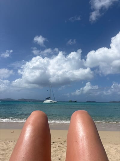 The scene depicts a first-person view from a reclining position on a sandy beach in Spanish Town, British Virgin Islands. In the immediate foreground, the tanned thighs and lower legs of a person are prominently featured, extending horizontally across the bottom of the frame. Beyond the sand, gentle waves lap at the shoreline, leading into clear, multi-toned turquoise and blue ocean water. In the mid-distance, a white catamaran sailboat with a visible mast and blue sail cover is anchored. To its right, a smaller, darker vessel is also present on the water. Distant, low-lying landmasses with green vegetation are visible on the horizon across the sea. The sky above is bright blue and occupies the upper half of the image, filled with numerous large, white cumulus clouds. The overall environment suggests a sunny day.