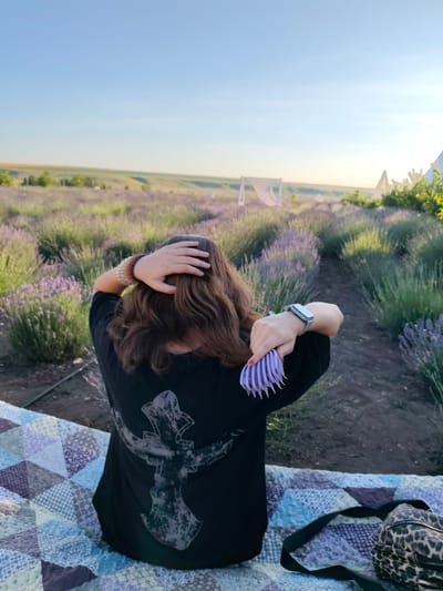A person sits on a quilt in a lavender field, brushing their hair. The person is wearing a black t-shirt with a large, faded graphic design on the back. They are also wearing a watch on their left wrist and several beaded bracelets on their right arm. The person is seated on a colorful patchwork quilt, and a leopard-print bag with a black strap rests beside them.
The setting is a vast lavender field, with rows of purple lavender stretching into the distance under a clear blue sky. In the background, there are several white tent-like structures and some trees. The sun appears to be low in the sky, suggesting either early morning or late afternoon, casting a warm light over the scene. The overall atmosphere is peaceful and serene.
The image was taken in Nechaiane, Ukraine.
