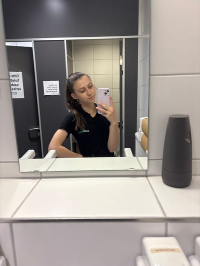 A woman is taking a mirror selfie in a restroom. She is wearing a black t-shirt and has long, dark hair. She is holding a light pink phone in her right hand. The restroom appears to be a public facility, with tiled walls and stalls visible in the reflection. There are signs in German on the stall doors. One sign reads "Bitte benutzen Sie den Papierhandtuchspender." Another sign is partially visible, but appears to be a general restroom instruction. The lighting suggests it is daytime.