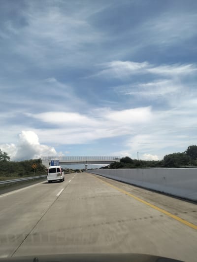 The image depicts a view from inside a vehicle traveling on a multi-lane highway in Keureuweungkrueng, Indonesia. The highway is paved with concrete and has clear yellow lane markings. On the right side of the highway, a tall, white concrete barrier runs along the edge. On the left side, there is a metal guardrail and a grassy embankment leading to dense green foliage and trees.
A white van is visible ahead in the left lane, moving in the same direction as the camera. Above the highway, a pedestrian overpass with a metal railing spans the road. Road signs are present near the overpass. One sign is blue and displays symbols indicating "P" (parking), a person with a bag, and "ATM". Another sign, yellow and diamond-shaped, shows a distance of "5.1 m".
The sky is a prominent feature, filled with dramatic, wispy white clouds against a bright blue background, suggesting a sunny day with some cloud cover. The lighting indicates it is daytime, likely late morning or early afternoon. The overall scene conveys a sense of travel and movement along a modern infrastructure route in a tropical or subtropical environment, characterized by lush vegetation under a dynamic sky. There is no explicit indication of people or specific emotional content beyond the context of a journey.