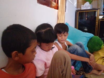 Three children are sitting together indoors. The child on the left wears an orange shirt, the middle child wears a pink shirt, and the child on the right wears a white tank top and blue jeans. They are sitting on a bed with blue and green pillows. A brown window frame is visible at the top of the frame on the left. Behind the children, on the right, is a dark brown cabinet with a tissue box on top. The location is Gampongjawa, Indonesia.