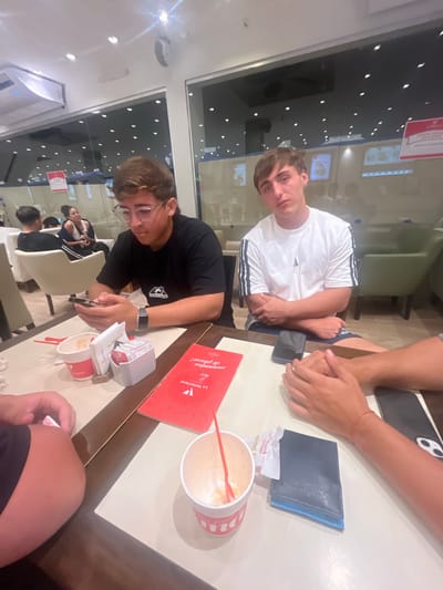 A high-angle indoor shot shows three young males seated at a table in what appears to be an ice cream parlor in Lanús Oeste, Argentina. The male at the top wears a white shirt with black stripes. The second male is in a black t-shirt and is focused on a cell phone in his hand. An ice cream container, several napkins, a wallet, and a menu are on the table, indicating the group is or has been eating. A second person's arm and phone are visible in the top right corner. The third individual is partially visible. The setting is indoors with reflective windows, hinting at a daytime scene. Chairs are also visible in the background, suggesting the dining area.