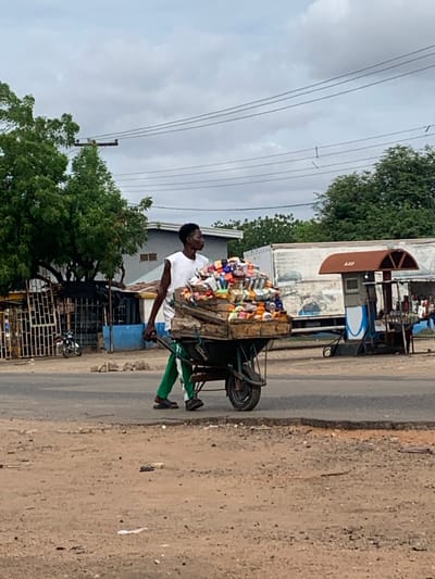An elevated, medium-shot photograph captured in Shendam, Nigeria, depicts a male street vendor pushing a heavily laden, two-wheeled cart from right to left across the foreground. The individual is a dark-skinned male, appearing young adult, wearing a white sleeveless undershirt, green track pants with a white side stripe, and dark sandals. He holds the cart's right handle with his right hand and looks towards the right. The cart, constructed with a wooden bed over a dark green metal frame, is piled high with a diverse assortment of small, colorful packaged goods, consistent with toiletries, cosmetics, or household items.
The scene takes place on the dirt shoulder adjacent to an asphalt road, which shows minor deterioration at its edge. The foreground is unpaved, dusty ground with scattered small stones and debris. In the mid-ground, a paved road stretches horizontally.
The background to the left features dense green trees partially obscuring various structures, including a grey building and a dark-colored motorcycle parked behind a white, gate-like fence structure. Further back, a small structure painted blue is visible. To the right background, beyond the paved road, a large white truck with faded graphics is parked. Adjacent to the truck stands an informal roadside stall characterized by a rusty-brown arched roof with "A & O" faintly visible, and a small enclosure. Bottles, presumably containing fuel, are arranged on a stand next to this structure, indicating a small-scale fuel sales point.
Overhead, an overcast sky with grey and white clouds dominates the upper frame. Multiple dark power lines traverse horizontally across the sky, supported by a wooden utility pole with crossbars visible through the trees on the left.