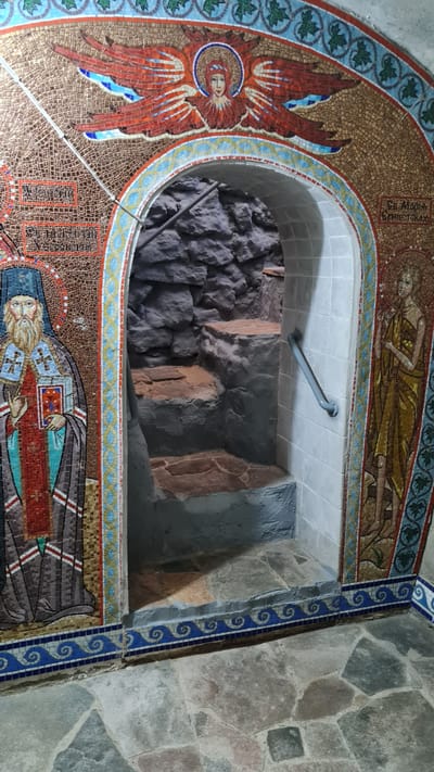 The image shows an interior view of a religious site with mosaic decorations. The foreground displays a rough stone floor. A mosaic archway, adorned with intricate patterns and depictions of religious figures and symbols, leads to a lower level. The mosaic on the left depicts a bearded male figure in religious attire holding a book. The mosaic on the right shows a female figure in a golden robe. Beyond the archway, a rough stone wall forms the background, with stone steps descending into a dimly lit area. A metal handrail is attached to the white tiled wall to the right of the archway. The overall scene suggests a historical or religious site, possibly a baptistery or a chapel, located in Bilhorod-Dnistrovskyi, Ukraine.