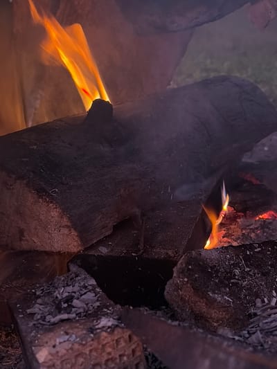 The image depicts a close-up view of a campfire. Several logs of wood are stacked, with flames and embers visible. The fire is burning on the logs, casting a warm glow. Smoke is also present, rising from the fire and obscuring parts of the background. The ground around the fire appears to be covered in ash and charred wood. The scene is dark, with the fire being the primary source of light. There are no people or animals visible in the image. The environment appears to be outdoors, with hints of greenery in the background, suggesting a natural setting.