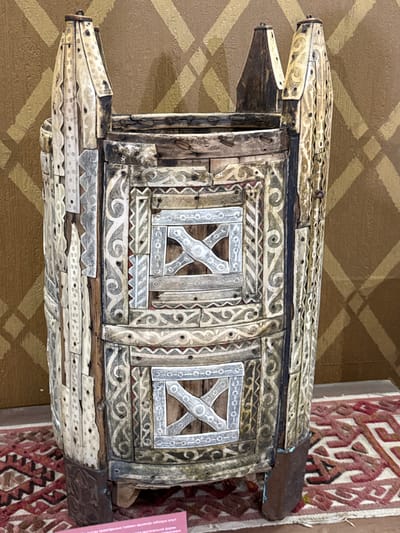 The image shows a close-up, eye-level, full shot of an intricately decorated wooden object. This object is standing on a rug with a red and white geometric pattern. The object itself is cylindrical with a flared top, appearing to be a type of container or furniture. It is adorned with numerous pieces of bone or ivory, carved with detailed patterns, which are affixed to the wooden structure with small nails or pins. The bone decorations include intricate scrollwork, geometric shapes, and circular motifs. The wooden parts of the object are weathered and show signs of age. The background consists of a wall with a repeating diamond-shaped pattern in gold and brown tones. A small pink label with text is visible at the bottom of the image. The object is located in the Museum of Fort-Shevchenko in Fort-Şevtşenko, Kazakhstan.