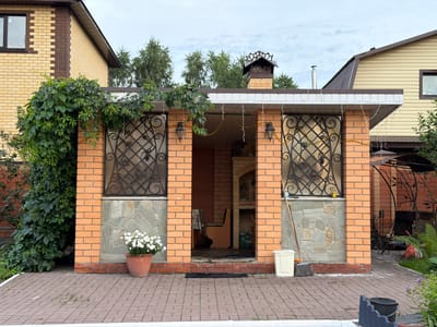 The image shows an outdoor brick structure, likely a gazebo or outdoor kitchen, in a residential setting. The structure is made of light orange bricks and has a white roof with decorative trim. Two windows with ornate black metal grates are visible on the front. A large vine with green leaves grows on the left side of the structure. White string lights are draped across the front of the roof.
In front of the structure, a paved patio area is laid with reddish-brown bricks. A terracotta pot filled with white flowers sits on the patio to the left of the entrance. The entrance itself is an open archway leading into the structure. Inside, a wooden bench and a brick fireplace or grill area are visible. To the right of the entrance, a broom and a white trash can are placed on the patio.
In the background, two houses are visible, one to the left and one to the right, constructed with yellow siding and brown accents. Trees with green foliage surround the area. The sky is overcast, suggesting a cloudy day. The overall scene depicts a well-maintained outdoor space in a suburban or rural environment. The location is identified as Kamenka, Russia. There is no discernible text in the image.