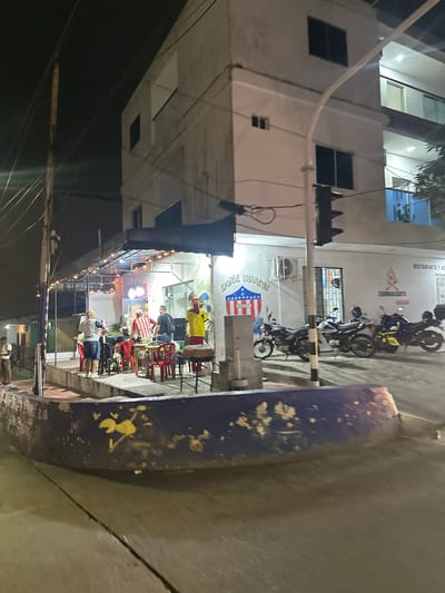 This is a nighttime street-level photograph of a restaurant and residential building in Perímetro Urbano Barranquilla, Colombia. In the foreground, a curved concrete barrier with faded yellow paint markings runs along the sidewalk. Beyond the barrier, the outdoor seating area of a restaurant is visible, with several tables and red chairs. Some people are seated at the tables, and others are standing. One man in a yellow shirt is standing near a grill. A sign on the building above the restaurant reads "DONDE IRRACOM JUNIOR." To the right of the restaurant entrance, a sign indicates "RESTAURANTE Y ASADOS CHURRASQUITO." Several motorcycles are parked in front of the building. The building itself is a multi-story structure with balconies and windows. The scene is illuminated by artificial lighting, creating shadows and highlights. Overhead power lines are visible against the dark night sky.