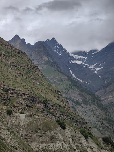 The image shows a mountainous landscape under a cloudy sky, likely in the vicinity of Gondhla, India. In the foreground, a steep, rocky mountainside covered with sparse vegetation slopes downwards from the left. A concrete structure resembling a staircase or retaining wall is visible on this slope. Behind this, a more distant mountain range rises, featuring sharp, rugged peaks. Some of these peaks are capped with snow, and a glacier is visible in a high mountain valley. The overall environment is one of dramatic, natural beauty characteristic of high-altitude regions.