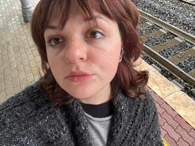 A close-up selfie-style photograph captures a single female individual on a train station platform. The subject, with light reddish-brown hair and bangs, brown eyes, and medium skin tone, appears to be in her 20s or 30s. She wears a small ring in her left nostril and a black earbud in her right ear. Her attire consists of a dark grey, open-knit cardigan over a light grey shirt, with a black collar visible underneath. She maintains a neutral expression, looking directly into the camera.

The background shows elements of a train station. To the subject's right, a set of railway tracks with metal rails, sleepers, and dark gravel ballast is visible. The platform itself features two distinct paving types: a light grey interlocking block pattern to the left, and a section of reddish-brown and beige patterned tiles with a partial yellow tactile strip along the platform's edge near the tracks. A dark metal post or lamp base is partially visible on the far left. The lighting is consistent with an overcast outdoor environment. This setting is identifiable as a train station, such as the one in Camucia, serving Cortona, Italy.
