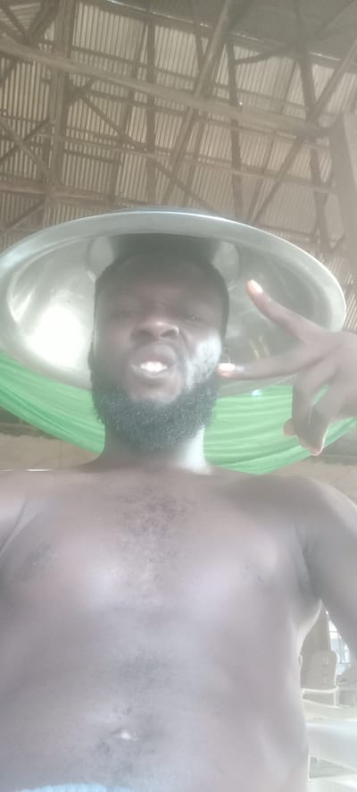A shirtless, dark-skinned man with a beard and a metal bowl on his head is posing for a selfie. He is making a peace sign with his right hand and grimacing. In the background, there is a green hammock and a corrugated metal roof supported by wooden beams. The image was likely taken in a building in Bende, Nigeria.