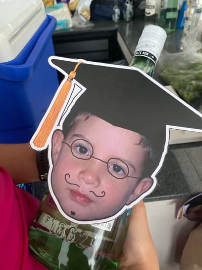 A person is holding a green glass bottle of Bacardi White Rum. A cutout image of a child wearing a mortarboard and drawn-on spectacles and mustache is superimposed on the bottle. A blue and white cooler sits on the left side of the frame. The setting appears to be indoors. There is no specific information in the image to confirm it is in Santa Cruz Tehuixpango, Mexico.