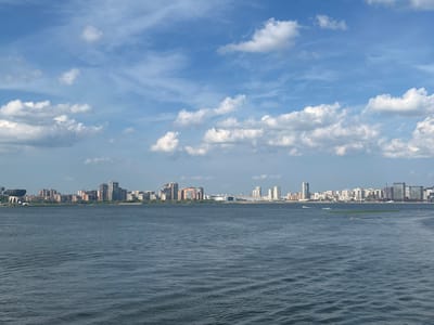 The image shows a panoramic view of the city of Kazan, Russia, as seen from the water. The foreground is dominated by a large body of water with ripples on its surface. In the middle ground, a city skyline stretches across the horizon, featuring a variety of buildings including residential high-rises, office buildings, and what appears to be a stadium or large circular structure on the left side. The buildings vary in height and architectural style, with some made of brick and others of glass and steel. The background consists of a bright blue sky with scattered white clouds. There are several small boats visible on the water, and a few boats and what appears to be a jet ski are also visible near the shoreline. The overall scene depicts a sunny day in a city located on a large body of water.