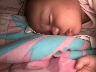 A close-up, indoor photograph shows a young infant asleep, nestled in a bed with a blanket. The infant's face, with closed eyes and slightly parted lips, is illuminated from the side, highlighting soft features and a few wisps of dark hair. The blanket is a prominent element, featuring a bold pattern of diagonal stripes in alternating shades of pink and teal. The fabric appears soft and quilted, suggesting a comfortable sleeping surface. A small portion of a cartoon-like image, possibly from a pillow or sheet, is visible in the upper left corner, featuring a simple face with two curved lines for eyes and a small circle for a mouth. The overall scene evokes a sense of peace and domesticity. The image was taken in Geulanggang Teungoh, Indonesia.