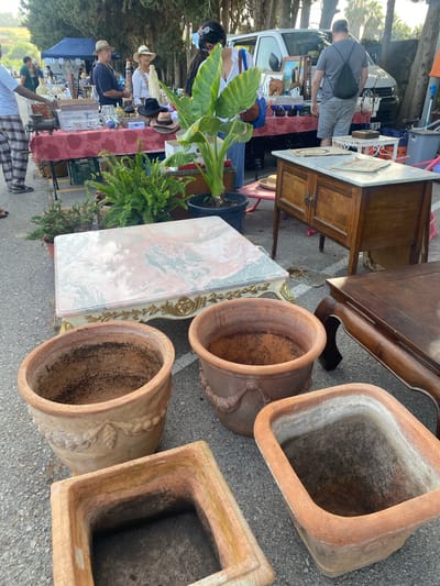 This image depicts an outdoor flea market or street sale, likely in San Luis De Sabinillas, Spain, given the presence of tall trees and what appears to be a Mediterranean climate. The scene is dominated by various items for sale, with a focus on furniture and decorative objects.

In the foreground, several large terracotta pots are displayed on the asphalt ground. One is a tall, rounded pot with a decorative rim featuring a leaf and vine motif. Next to it, a square, shallow terracotta planter is visible. Another rounded pot, larger and more ornate than the first, is positioned to the right.

Behind the pots, a rectangular coffee table with a pink and green marble top rests on a white ornate base with gold detailing. To the right of this coffee table, a dark wooden coffee table with carved legs is present. Further back, a small wooden cabinet with two doors and a marble top is displayed, with papers on top.

In the middle ground, a vendor's table is covered with a red and white patterned cloth. On this table are various items including what appears to be a meat grinder, a white decorative metal piece, a stack of books or magazines, and several hats. A large green potted plant with broad leaves stands near the table.

Several people are visible in the background, engaged in browsing or selling. A man in a grey t-shirt and shorts, wearing a backpack, is looking at items on a table. A woman in a white shirt and hat, and a man in a blue shirt and hat, are also present near a stall. A blue tent is visible in the far background. The overall setting suggests a sunny day at a bustling outdoor market.