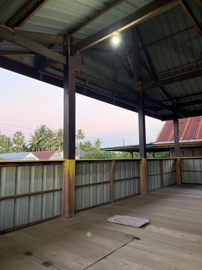 The image depicts a covered wooden porch or open-air structure with a corrugated metal roof supported by wooden beams and posts. A single lightbulb illuminates the interior. The porch overlooks a landscape of palm trees and buildings with metal roofs. In the foreground, a fluffy, light-colored mat is placed on the wooden floor. The scene appears to be located in Turumame, Indonesia, as suggested by the presence of palm trees and the style of the buildings. The sky is a soft gradient of pink and blue, indicating either sunrise or sunset.