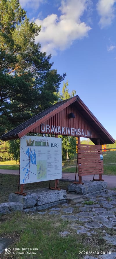 This media file captures a sign for "ORAVAKIVENSALIM" and "HARTOLA INFO" in Joutsa, Finland. The sign is wooden and features silver lettering. To the right of the sign, there is a wooden slatted information board, and to the left, a tall pine tree. The ground is covered in stones and grass. The sky is blue with some white clouds, suggesting it is daytime with fair weather. The timestamp on the image indicates it was taken on June 30, 2026, at 20:18, suggesting it's early evening during the summer. The text on the sign, "Yrittäjäystävällinen Hartola" and "www.hatola.fi," indicates that Hartola is a business-friendly municipality. The scene appears to be an outdoor information point or entrance to a recreational area. The presence of the camera's watermark, "REDMI NOTE 9 PRO AI QUAD CAMERA," is also visible. There are no people visible in the image.
