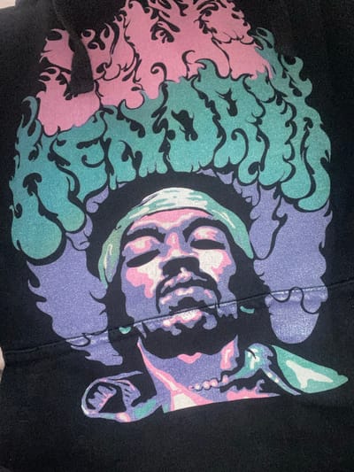 A close-up image displays a vibrant, multi-colored graphic design printed on black fabric, likely part of a garment. The central element of the design is a highly stylized, abstract face or skull presented in profile, facing towards the right. This face is rendered in shades of lavender, pastel pink, and white, with subtle light green accents, and features prominent eye sockets, a defined nose area, and an open mouth. Surrounding the face are dynamic, swirling patterns reminiscent of clouds, flames, or intricate natural forms. These patterns are depicted in soft pink and a shimmering teal or mint green, with distinct black outlines that enhance their complex shapes. A visible vertical seam or fold runs down the center of the graphic, passing through the stylized face. The print itself exhibits a slightly textured, granular quality, characteristic of screen printing. The overall aesthetic is abstract and fantastical, utilizing a palette of bright, contrasting pastel colors against the deep black background. No discernible activity is taking place, as it is a static image of a design. There is no visible text.