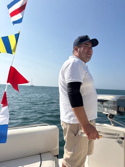 A man wearing a dark blue baseball cap with an anchor emblem, a white polo shirt, and khaki shorts stands on the deck of a boat. He is holding a pair of stainless steel tongs with wooden handles. To his left, nautical flags are visible, including a red pennant flag and a flag with blue and yellow stripes. In the background, the ocean is calm, and a sailboat is visible in the distance. The scene is likely taking place in the waters off Callao, Peru, a major port city known for its maritime activities.