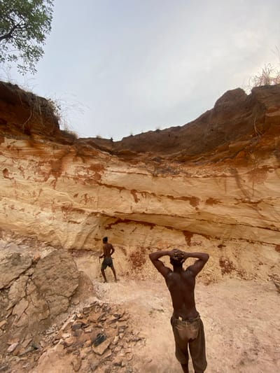 The image depicts an open excavation site in Amoso, Nigeria, under an overcast sky. A large, stratified earthen and rock face dominates the background, exhibiting distinct horizontal layers of light beige material interspersed with reddish-brown streaks and darker brown soil at the top. Sparse dry grass and a leafy tree branch are visible along the upper edge of the excavation.
Two shirtless adult males are present within the excavation. In the foreground, slightly right of center, a man with dark skin is seen from the back, wearing light-colored, patterned trousers or long shorts. His hands are clasped behind his head in a posture of rest or observation. Further back and to the left, a second man with dark skin is partially visible, facing towards the rock face. He wears dark shorts and holds what appears to be a pickaxe or similar digging tool, resting its head near the ground. The ground beneath the men is uneven, composed of loose reddish-brown soil, sand, and piles of rock fragments, particularly evident in the mid-ground. The overall scene suggests manual labor within a quarry or mining operation.
