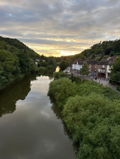 This image depicts a view of the River Severn in Ironbridge, Shropshire, United Kingdom, at dusk. The sky is overcast with patches of sunlight breaking through, casting a warm glow on the scene. On the right bank of the river, a row of historic buildings, typical of a Georgian or Victorian style, lines a street. These buildings appear to be shops and residences, with visible windows, doors, and chimneys. Cars are parked along the street, and a few people can be seen walking. The river itself is calm and reflects the sky and surrounding trees. The left bank of the river is densely wooded with lush green foliage, creating a natural contrast to the built-up area on the opposite side. The overall atmosphere is peaceful and picturesque, capturing the charm of this historic town.