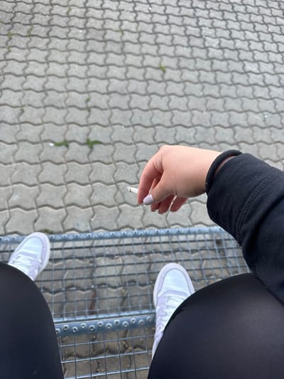 An individual, likely female based on the hand shape and nail polish, is seated outdoors on a metal bench. They are wearing black leggings and white sneakers. The individual is holding a lit cigarette between their index and middle fingers. The background consists of interlocking grey pavers with scattered green weeds and a slightly blurred metal grating beneath the bench. The scene appears to be in a public outdoor space, possibly a park or seating area in the city of Bad Säckingen, Germany.