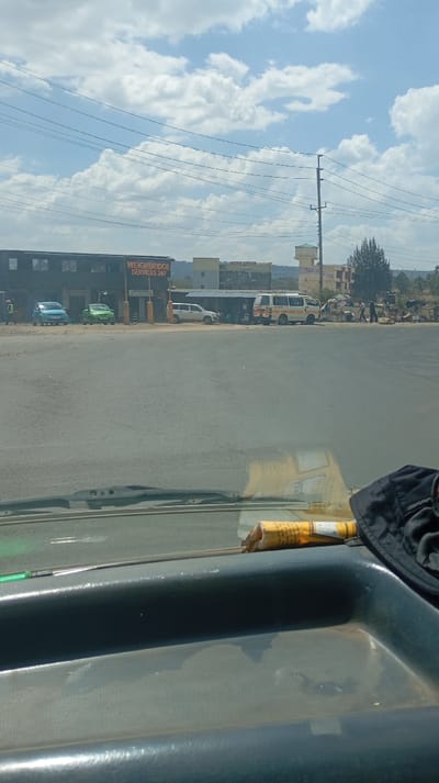 A street view features "WEIGHBRIDGE SERVICES 24/7" signage on a building, with cars parked nearby. A white van and a bus are farther down the road, where people gather. Overhead power lines crisscross the sky, dotted with fluffy white clouds. The foreground is dominated by a car dashboard holding a rolled-up paper and a black hat.