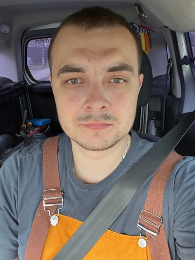 The image shows a young man positioned diagonally across the frame, his head towards the upper left, appearing to take a selfie or looking into the camera while seated in a vehicle. He has short dark hair, light stubble on his chin and upper lip, and light brown or hazel eyes looking directly forward. He is wearing a dark grey t-shirt and a silver chain necklace. Over his t-shirt, he wears an orange garment, possibly an apron or overalls, secured by a brown fabric strap with a rectangular metal buckle. A black car seatbelt crosses his chest.
The background reveals the interior of a vehicle, likely a car or van. To the upper left, behind his head, a stack of colorful folded items (red, white, blue, yellow, orange) is visible. Towards the lower right, behind his shoulder, a blue toolbox and various tools, including pliers and wrenches, are visible on the floor, alongside a black plastic bag. Through a small back window, faint reflections or patterns of a building exterior with vertical lines can be seen, along with some indistinct text that appears to include "ANH". On the silver button of the brown strap, the word "JEANS" is visible, surrounded by small stars. The man has a neutral to slightly serious expression. The lighting suggests it is daytime.