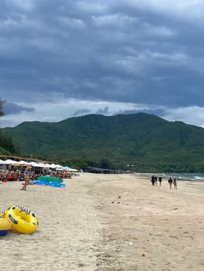 The image depicts a wide, sandy beach in Cam Lam, Vietnam, under a cloudy sky. In the foreground, two inflatable pool rings, one yellow with blue accents and "Perfect" written on it, and another partially visible yellow and blue ball, rest on the sand. A woman in a sunhat and yellow bikini walks away from the viewer towards the left. Behind her, a row of lounge chairs with people sitting and lying on them are set up under numerous white umbrellas. Several inflatable dolphin floats and a green surfboard are also present on the sand. Further back, a line of small structures with thatched roofs and more umbrellas marks the edge of the beach. In the middle ground, a group of people walks along the shoreline. In the background, a densely forested, green mountain rises dramatically. The sea is visible on the right side of the image, with small waves and a few people in the water. The overall atmosphere is one of a beach resort on a slightly overcast day.