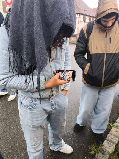 The image depicts three individuals outdoors on a paved surface in Meistratzheim, France, under overcast lighting conditions. In the foreground, an individual with dark, curly hair, partially obscured by a dark grey fringed scarf draped over their head and shoulders, wears a light blue denim jacket, light blue denim jeans, and white athletic shoes. This individual's right hand holds a black smartphone horizontally, displaying an indistinct image or video, while their left hand is in the front pocket of their jeans. To the right, a second individual, a male, wears a two-tone tan and black hooded jacket with the hood up, light blue loose-fitting jeans, and dark-colored shoes. He carries a black backpack and has both hands in his jacket pockets, with his head tilted downwards. A third individual's lower leg and white shoe are partially visible on the far left. The ground consists of a dark, wet paved surface, with a narrow strip of cobblestone and sparse green vegetation along the right edge. In the background, a traditional half-timbered house with a red tiled roof is visible, characteristic of the Alsace region.
