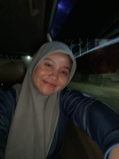 The image is a close-up, vertically oriented selfie of a young woman wearing a grey hijab and a dark blue shirt. Her face is the primary subject, with her head tilted and a slight smile on her face. The background is dark and indistinct, with hints of blurry lights and structures, suggesting an indoor or nighttime outdoor setting. The lighting is somewhat dim, with a slight sheen on her skin, possibly from makeup or ambient light. One of the crops shows a faint hint of what might be a sign or poster with colorful elements in the dark background. The overall mood appears casual and relaxed. The image was taken in Sungai Paring, Indonesia.