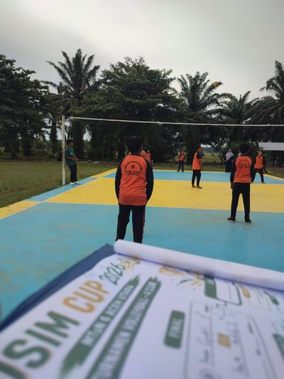 The image depicts an outdoor volleyball game in progress on a court with blue and yellow markings, located in Blangriek, Indonesia. Several young males, dressed in athletic attire, are actively participating in the game. Some are positioned on the court, seemingly preparing to receive or serve the ball, while others are in the background, observing or waiting for their turn. A yellow volleyball is visible in the air near a palm tree. The environment is characterized by lush green trees, including prominent palm trees, and a cloudy sky overhead. In the foreground, a clipboard with a document is partially visible, suggesting that the game might be part of an organized event. The overall scene conveys a sense of youth sporting activity in a tropical setting.