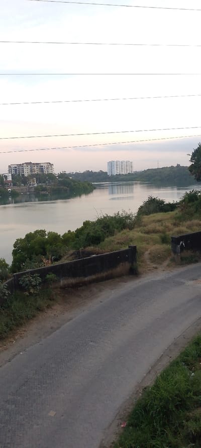 The scene depicts a panoramic view of a waterway in Junda ward, Kenya, with urban structures on the far bank. In the foreground, a dark grey paved road, constructed of interlocking pavers, slopes diagonally from lower left to middle right. A light brown rectangular object lies flat on the road near its center. To the immediate right of the road is a strip of green grass and dirt with minor debris. To the left, a dark grey concrete retaining wall or barrier runs parallel to the road, featuring a central opening that reveals a dirt path leading uphill. Beyond this wall and along the near bank of the waterway is dense green vegetation consisting of shrubs and trees.
The midground is dominated by a calm body of water, likely a lake or wide river, which reflects the overcast sky and distant structures. Lush green foliage covers the banks bordering the water. In the background, on the far bank, two prominent multi-story buildings are visible. To the left, a lighter-colored building with multiple balconies and a darker roofline stands amidst trees. Further to the right, a taller, white, multi-story residential or commercial building is visible, with its reflection clearly cast upon the water's surface. The sky is a pale grey-white with faint clouds. Multiple thin, dark utility wires are stretched horizontally across the upper third of the image. No individuals or active vehicles are present.