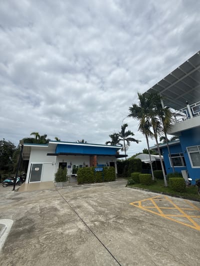 The image shows the exterior of a building with a blue and white facade, located in Don Sak, Thailand. The building appears to be a commercial establishment, possibly a restaurant or a shop, with a sign indicating "WOMEN". In front of the building, there is a paved area with a yellow star-shaped marking. Palm trees are visible in the foreground and background, suggesting a tropical environment. The sky is overcast with clouds. There are no people visible in the image.