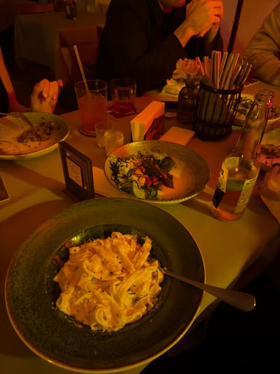 The image shows a dinner table with plates of food, glasses of drinks, a bottle, and cutlery in ქვედა სამება, Georgia. A large plate of fettuccine pasta with a creamy sauce is visible. There's also a bowl of salad. Several people are seated around the table, with their hands and arms visible. The lighting is dim and warm, possibly from artificial light sources, creating a cozy atmosphere.