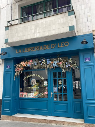 The image displays the teal-blue storefront of "LA LIBRERÍA DE Dª LEO," a bookstore located at number 21 in Buñol, Spain. The name of the shop is rendered in raised gold letters above the main facade. Two black, industrial-style lamps are mounted symmetrically on the blue facade. A vibrant, ornate floral arrangement of pink, blue, and white flowers, interspersed with dried foliage, drapes across the top of the large display window and the double entrance door.
The left side of the storefront features a display window showcasing various books, including one by Máximo Huerta. A prominent circular logo depicting a white dog wearing glasses, with "LA LIBRERÍA DE DOÑA LEO" inscribed around it, is visible on the window glass. A small, light-colored dog figure is positioned in the lower-left corner of this window. The entrance, a double door with glass panes on the left leaf, displays a "CERRADO" sign and other informational stickers.
Above the storefront, a balcony with a rusty metal railing extends from the building. The wall directly above the bookstore's sign and below the balcony is faced with light-colored square tiles, accented by intermittent teal tiles. On the floor above the balcony, three green-framed sliding windows are visible, behind which purple sheer curtains are drawn. White conduits or pipes run vertically on the left side of the building. The storefront opens directly onto a light-colored paved area, under an overcast sky. No individuals are present in the scene.