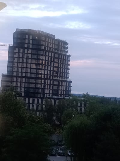 The image shows a tall, modern building with many windows, suggesting it is an apartment complex or office building. The building is dark in color, with lighter vertical elements. The top of the building has a sign that reads "TRUST INVESTMENT". The building is surrounded by trees, and in the foreground, there are cars parked in a lot. The sky is cloudy and appears to be dusk or dawn. This building is located in Kielce, Poland, and is known as "Budynek Alfa".