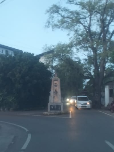 This image depicts a street scene in Meunasah Papeun, Indonesia, likely captured during the early evening or twilight given the dim lighting and illuminated headlights of vehicles. The central subject is a tall, weathered monument or obelisk with inscriptions, positioned at what appears to be a junction or intersection. Flanking the monument are dense trees and foliage, with buildings visible in the background, including one with a multi-story structure.

Two cars are on the road, with their headlights on, suggesting low light conditions. One is a white van, and another vehicle is partially visible behind it. The road has a curved white line marking, indicating a lane division or turn. The sky is a dusky blue. The overall atmosphere is calm and quiet, with no explicit signs of human activity beyond the vehicles and potentially distant figures on the right side of the road. The image is somewhat blurry, contributing to a slightly indistinct visual impression of the scene. No discernible text is readable on the monument or any other surfaces.