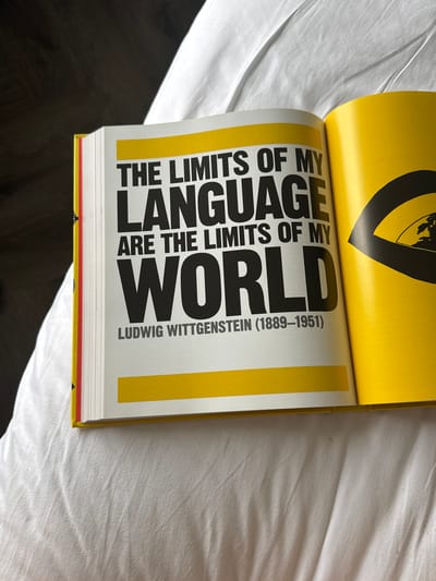 The image shows an open book with a quote by Ludwig Wittgenstein. The quote reads: "THE LIMITS OF MY LANGUAGE ARE THE LIMITS OF MY WORLD." Below the quote is Wittgenstein's name and dates: "LUDWIG WITTGENSTEIN (1889-1951)". The text is in a bold, black sans-serif font against a white background. The pages of the book have a yellow border at the top and bottom. The book is open on a white, rumpled surface that appears to be a bedspread or similar fabric. The background is out of focus and dark on the left, suggesting a dimly lit room. There is a partial view of another page on the right, which is solid yellow with a black graphic element. There are no people or animals in the image. The image does not provide any information about the location of the city of Da Loc, Vietnam.