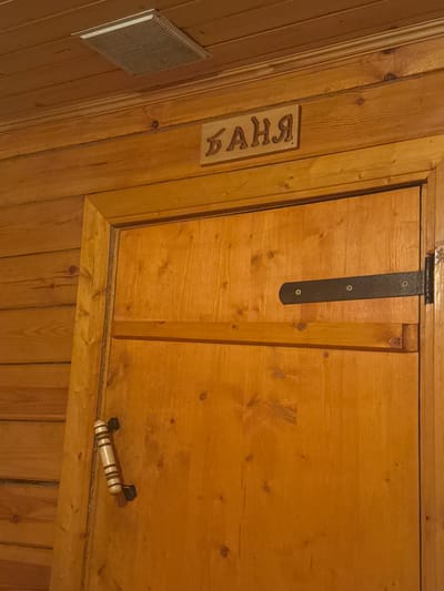 This is a medium-shot interior photograph taken at a close angle, showcasing a wooden door within a sauna. The door is made of light-colored wood and features a wooden handle and a black metal hinge. Above the door, a sign with Cyrillic lettering, reading "баня" (banya, meaning "bathhouse"), is affixed to the wooden wall. To the left of the sign, a ventilation grate is visible on the wooden ceiling. The walls and ceiling are constructed from horizontal wooden planks. The overall scene depicts a rustic and functional sauna interior. The location is identified as Mayak, Russia. There are no people or visible actions occurring in the image.