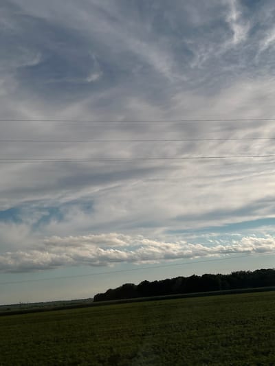 This forensic image captures a daytime rural landscape in Velyki Hai, Ukraine. The upper two-thirds of the frame feature a sky characterized by extensive, layered cloud formations, including wispy high-altitude cirrus and fragmented lower cumulus clouds, with intermittent patches of light blue sky visible predominantly towards the lower left. Four dark, parallel power lines are horizontally positioned across the mid-upper portion of the sky. A distinct, dark green tree line or forest forms a horizontal band across the mid-ground. The foreground consists of a large, uniformly green agricultural field with visible crop texture, extending towards the bottom edge of the frame. In the far left distance, subtle vertical structures, likely power pylons, are faintly visible against the horizon. No people, animals, or specific actions are observed.