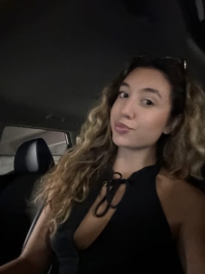 The media file is a low-angle, dimly lit selfie of a young woman with curly blonde hair, lying down and looking at the camera. She is wearing a black top with a laced neckline. The background is dark, suggesting an indoor setting or nighttime, possibly within a car given the curved dark surface on the left and the presence of a car mirror on the right. Her expression is relaxed and slightly playful. No text is visible. The overall mood is intimate and casual.
