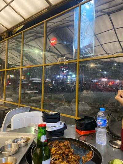 This is a medium shot photograph taken from inside a restaurant, looking outwards through a glass-paneled roof structure with a yellow metal frame. The perspective is slightly tilted, with the bottom left of the image showing a white corrugated roof or awning.
The interior of the restaurant is visible in the foreground, with a table set for dining. On the table, there are metal serving trays with small bowls, a green glass bottle with a red cap, and a black and orange plastic container. A partially eaten dish of meat is in a round black pan. A black handbag and a plastic water bottle with a blue cap are also on the table.
Through the glass, the exterior scene at night is visible. It appears to be a street with many motorcycles and cars. There are neon signs and streetlights, indicating a busy urban environment. One prominent sign visible through the glass is for "Richeese Factory," a fast-food chain. Another sign in the background reads "GALERY JAM TANGAN" (Watch Gallery) with an address and social media handle from Bandar Lampung, Indonesia. The overall atmosphere suggests a lively evening meal or gathering in an urban setting.