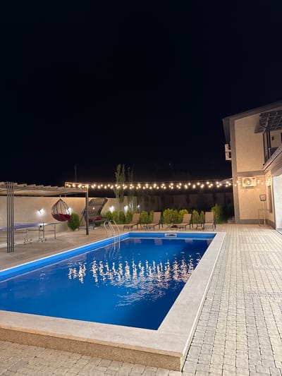 The image depicts an outdoor swimming pool area at night, illuminated by string lights and wall-mounted lamps. The pool, filled with bright blue water, has a reflection of the string lights on its surface. Surrounding the pool is a paved patio. To the left of the pool, there is a covered area with a ping pong table and a hanging chair. In the background, there are several lounge chairs, small trees, and a building. The scene is set in the city of Mughni, Armenia. There are no people or discernible actions visible in the image.